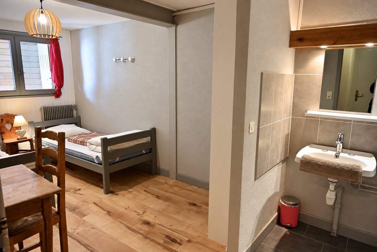 Chambre simple avec lit gris, parquet clair et lavabo